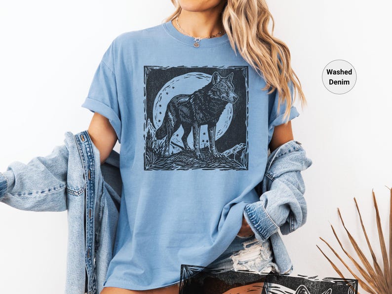 May include: Light blue t-shirt with a black and white wolf illustration. The wolf is standing in front of a full moon. The shirt is made of a soft material and has a relaxed fit. The words "Washed Denim" are printed on the shirt.