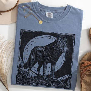 May include: A blue jean-colored t-shirt featuring a black wolf illustration against a moonlit background. The shirt is paired with brown sandals, a hat, and a gold necklace. The wolf design is detailed and artistic.