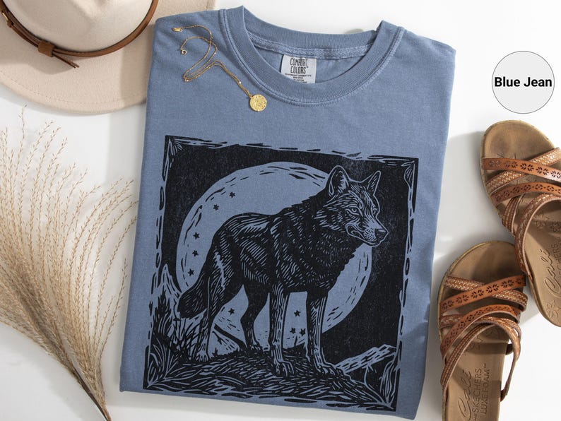 May include: A blue jean-colored t-shirt featuring a black wolf illustration against a moonlit background. The shirt is paired with brown sandals, a hat, and a gold necklace. The wolf design is detailed and artistic.