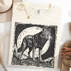 May include: Ivory-colored t-shirt featuring a black and white wolf illustration against a full moon and stars. The shirt is made of a soft material. A gold necklace and a beige hat are also in the image.