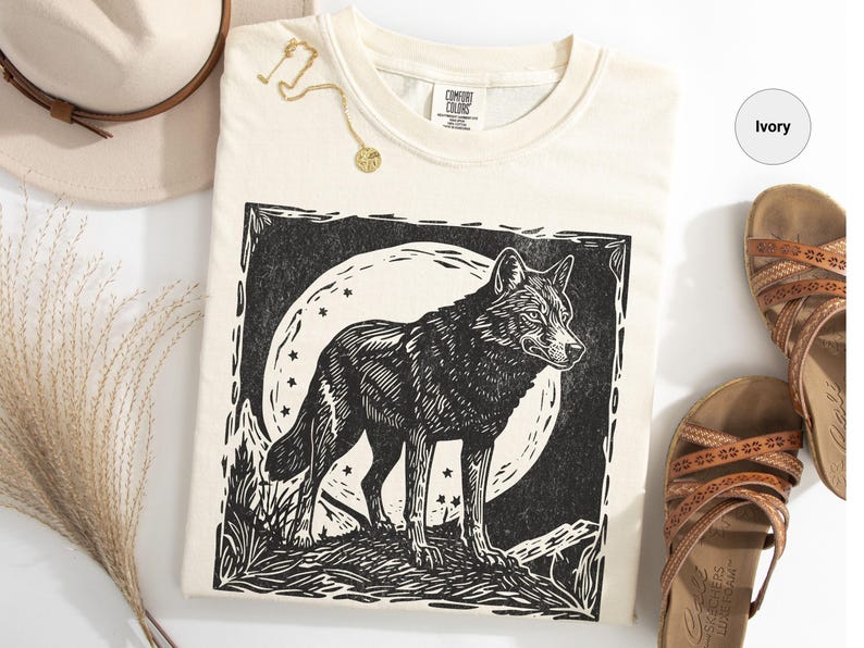 May include: Ivory-colored t-shirt featuring a black and white wolf illustration against a full moon and stars. The shirt is made of a soft material. A gold necklace and a beige hat are also in the image.