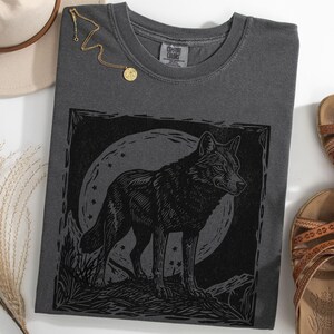 May include: Pepper-colored t-shirt featuring a black wolf illustration against a moonlit background. The shirt is paired with brown sandals, a gold necklace, and a beige hat. The overall theme is nature and wildlife.