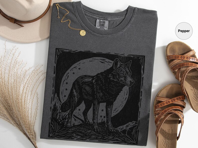 May include: Pepper-colored t-shirt featuring a black wolf illustration against a moonlit background. The shirt is paired with brown sandals, a gold necklace, and a beige hat. The overall theme is nature and wildlife.