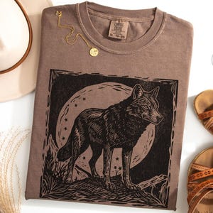 May include: Espresso-colored t-shirt with a black wolf graphic, a gold necklace, and brown sandals. The wolf design is set against a moon and stars. A beige hat is visible in the upper left corner.