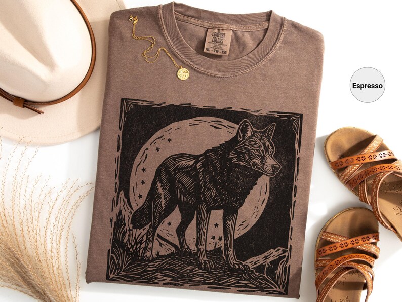 May include: Espresso-colored t-shirt with a black wolf graphic, a gold necklace, and brown sandals. The wolf design is set against a moon and stars. A beige hat is visible in the upper left corner.