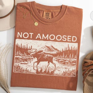 May include: A rust-colored t-shirt with the words "NOT AMOOSED" above a graphic of a moose in a river. The shirt is paired with a hat, sandals, and a gold necklace.