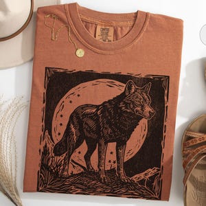 May include: A terracotta-colored t-shirt featuring a black graphic of a wolf standing before a full moon. A gold necklace and a beige hat are also visible. Brown sandals are placed to the right of the shirt.