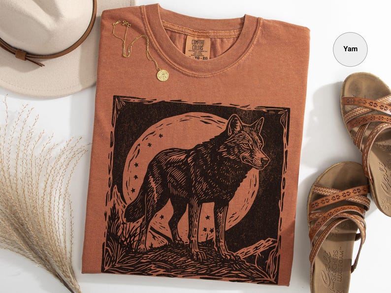May include: A terracotta-colored t-shirt featuring a black graphic of a wolf standing before a full moon. A gold necklace and a beige hat are also visible. Brown sandals are placed to the right of the shirt.