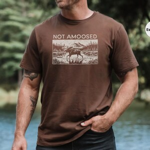 May include: Espresso-colored t-shirt featuring a rectangular graphic with a moose in a landscape, mountains, and trees. The text "NOT AMOOSED" is above the graphic. The shirt is short-sleeved.