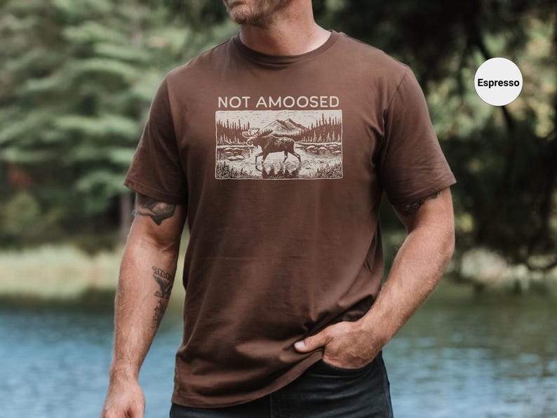 May include: Espresso-colored t-shirt featuring a rectangular graphic with a moose in a landscape, mountains, and trees. The text "NOT AMOOSED" is above the graphic. The shirt is short-sleeved.
