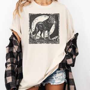 May include: Ivory-colored t-shirt featuring a black and white graphic of a wolf standing in front of a full moon. The wolf is detailed with a woodcut style. The shirt is worn with denim shorts and a black and white plaid shirt.
