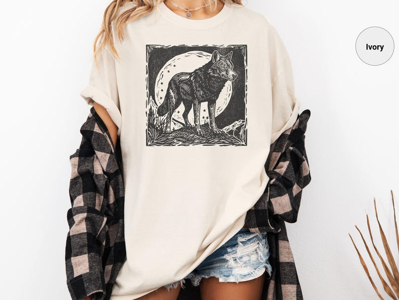 May include: Ivory-colored t-shirt featuring a black and white graphic of a wolf standing in front of a full moon. The wolf is detailed with a woodcut style. The shirt is worn with denim shorts and a black and white plaid shirt.