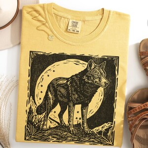 May include: Mustard-colored t-shirt with a black and white wolf graphic. The wolf is standing in front of a full moon with stars. The shirt is styled with brown sandals and a cream-colored hat.