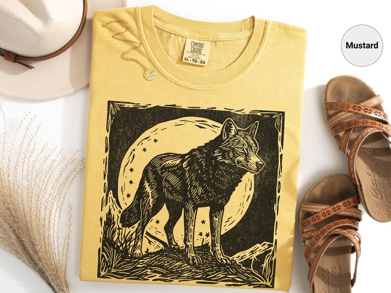 May include: Mustard-colored t-shirt with a black and white wolf graphic. The wolf is standing in front of a full moon with stars. The shirt is styled with brown sandals and a cream-colored hat.