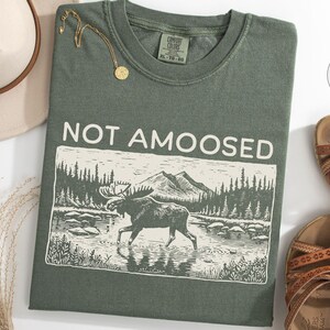 May include: Moss green t-shirt with the phrase "NOT AMOOSED" above a graphic of a moose in a river with mountains in the background. The shirt is next to a hat, sandals, and a gold necklace.