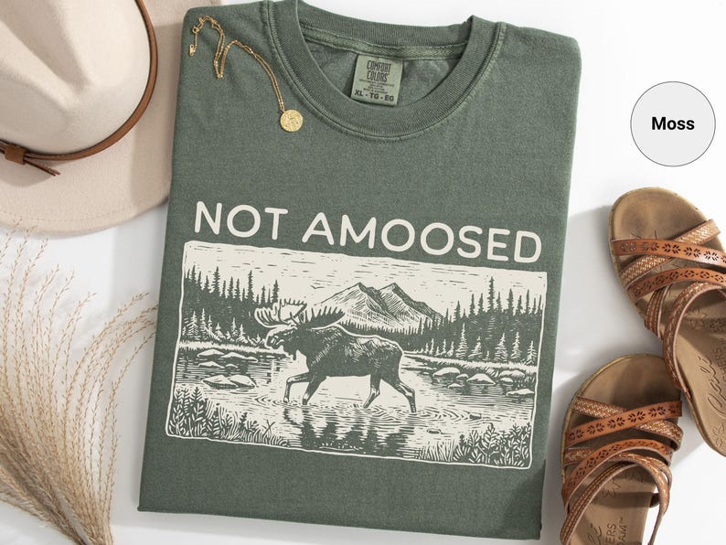 May include: Moss green t-shirt with the phrase "NOT AMOOSED" above a graphic of a moose in a river with mountains in the background. The shirt is next to a hat, sandals, and a gold necklace.
