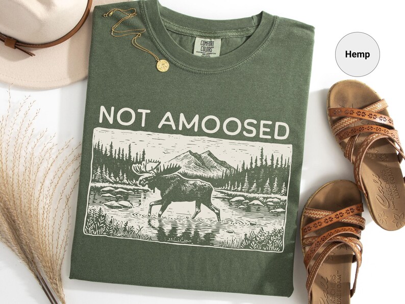 May include: Olive green t-shirt with the text "NOT AMOOSED" above a black and white illustration of a moose in a river. The shirt is paired with brown sandals, a gold necklace, and a beige hat.
