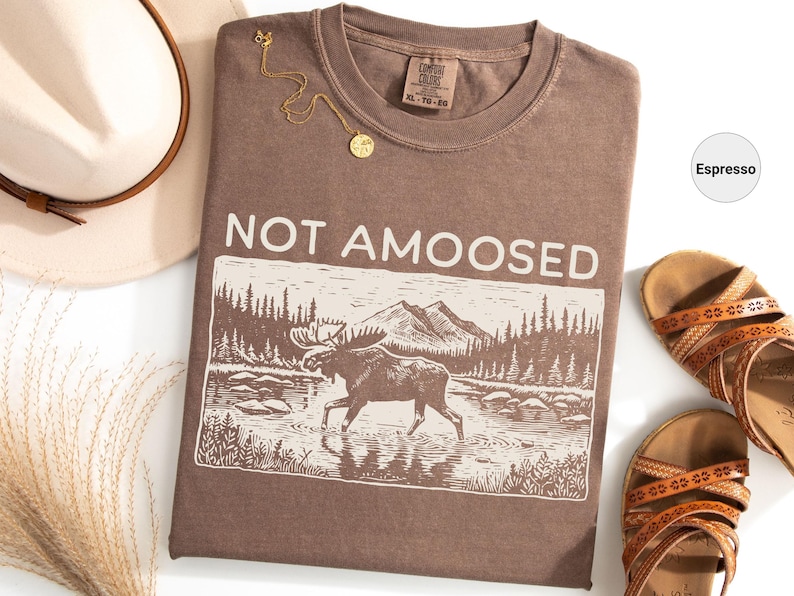 May include: Espresso-colored t-shirt with the text "NOT AMOOSED" above a graphic of a moose in a river. The shirt is paired with a hat, sandals, and a gold necklace. The shirt is a Comfort Colors XL-TG-2G.