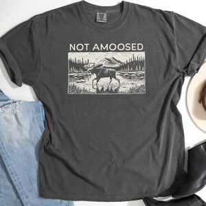 May include: Dark gray t-shirt with the text "NOT AMOOSED" above a black and white graphic of a moose in a lake. The shirt is paired with blue jeans, a white hat, and black boots.