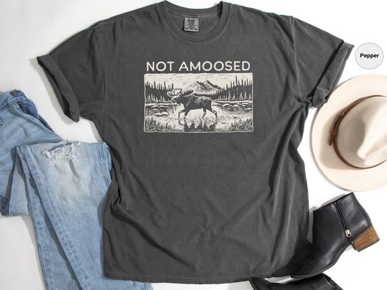 May include: Dark gray t-shirt with the text "NOT AMOOSED" above a black and white graphic of a moose in a lake. The shirt is paired with blue jeans, a white hat, and black boots.
