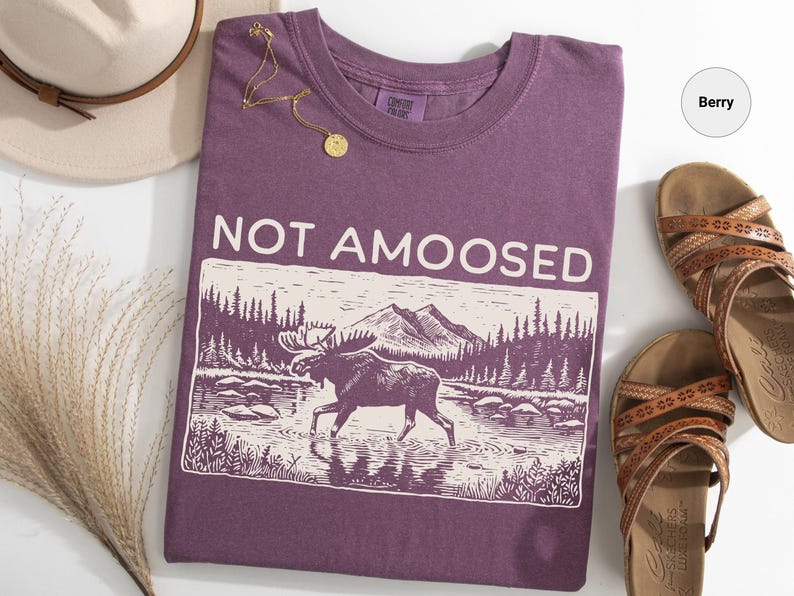 May include: Berry-colored t-shirt with the text "NOT AMOOSED" and a graphic of a moose in a river with mountains in the background. A gold necklace, a pair of brown sandals, and a beige hat are also in the image.
