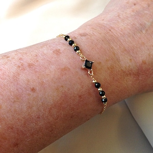 May include: A delicate gold bracelet with a central black square gemstone and small black beads. The bracelet is strung on a fine gold chain, creating a minimalist and elegant design.