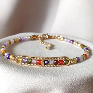 May include: A gold bracelet with a central rectangular section set with colourful gemstones. The bracelet's band is made of small, multi-coloured beads. The clasp and connecting chain are also gold, creating a delicate and elegant piece of jewellery.