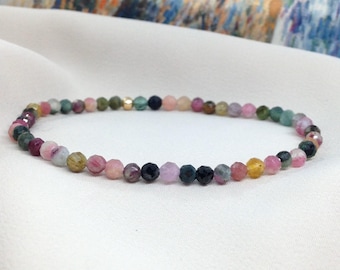 May include: A delicate bracelet featuring small, faceted beads in a variety of colors including pink, green, yellow, and black. The bracelet has a gold-colored accent bead. The beads are strung together to form a complete circle.