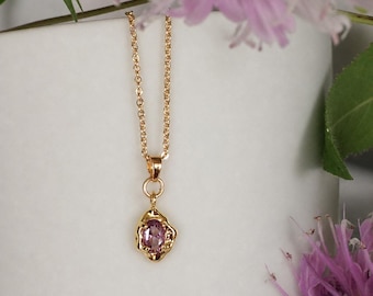 May include: A gold chain necklace featuring a small, oval-shaped pink gemstone pendant. The pendant is set within a textured, irregular gold frame that mimics a molten or organic metal finish, suspended from a delicate gold link chain.