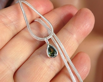 May include: A silver necklace with a teardrop-shaped pendant. The pendant features a dark green gemstone set in a silver bezel. The chain is a delicate, woven design. The necklace is displayed against a neutral background.