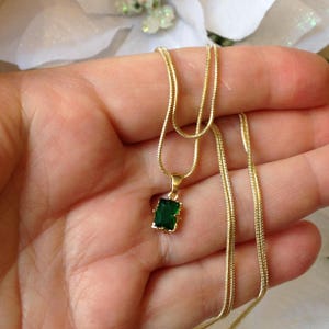 May include: A gold-colored necklace with a rectangular emerald-green pendant. The pendant is set in a gold frame and hangs from a delicate gold chain. The necklace is displayed against a soft, blurred background.