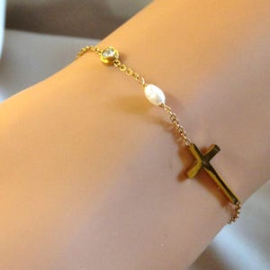 May include: Gold bracelet with a cross pendant, a small clear gemstone, and a white pearl. The fine chain bracelet is designed with a classic and elegant style.