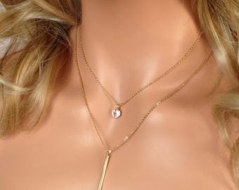 May include: A layered gold necklace with two strands. The top strand features a small, round, clear gemstone pendant. The bottom strand has a vertical bar pendant. The necklace is displayed on a person's neck.
