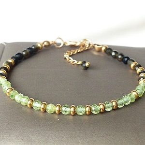 May include: A delicate bracelet featuring a combination of light green and dark green beads, interspersed with small gold-colored beads. The bracelet has a gold-colored chain and clasp. The beads are small and faceted, reflecting light. The bracelet is designed for a comfortable fit.