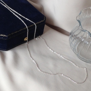 May include: A delicate silver chain necklace with a small gold pendant. The necklace is displayed on a white surface next to a dark blue jewelry box and a clear glass vase. The chain is approximately 50 cm long.