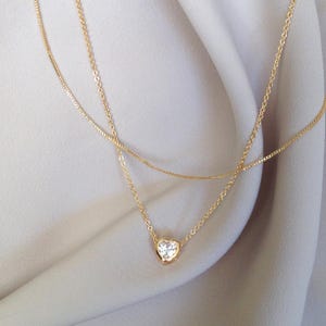 May include: A delicate gold necklace with two strands. The lower strand features a heart-shaped pendant with a clear, sparkling stone. The necklace is displayed against a soft, neutral-coloured fabric.