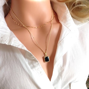 Peut inclure: Un collier doré à plusieurs rangs avec un pendentif noir rectangulaire. Le pendentif est encadré de petites pierres transparentes. Le collier est présenté sur une chemise blanche.