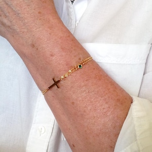 May include: A gold-coloured bracelet with a cross pendant and a small green gemstone. The fine chain bracelet is worn on an arm, set against a white shirt. The cross is a key design feature.