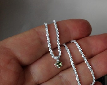 May include: A delicate silver necklace with a small, round, green gemstone pendant. The chain is made of tiny, sparkling silver beads. The necklace is displayed on a neutral background, highlighting its elegant design.