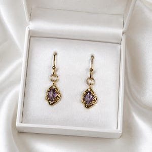 Peut inclure: Une paire de boucles d'oreilles pendantes dorées avec des pierres violettes à facettes de forme ovale, serties dans des montures en métal texturé irrégulier. Chaque boucle est suspendue à un crochet doré, présentée dans un écrin blanc satiné.