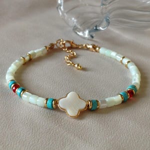 Peut inclure: Un bracelet délicat avec un charm en forme de trèfle blanc bordé d'or. Le bracelet est orné de perles blanches, turquoise et rouges, et d'accents dorés. Le fermoir est doré.