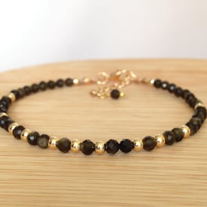 May include: A delicate bracelet featuring faceted black beads and gold-coloured metallic beads. The bracelet is strung on a gold-coloured chain with an adjustable clasp. The jewellery is displayed on a light brown wooden surface.