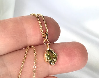 May include: A gold-toned necklace featuring a delicate chain and a small, irregular-shaped pendant with a faceted olive-green stone set in a gold-coloured metal frame. The pendant hangs from a simple jump ring attached to the fine link chain.