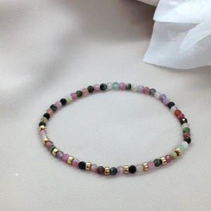 Peut inclure: Un bracelet délicat orné de petites perles de pierres précieuses multicolores dans les tons de rose, vert, noir et or. Le bracelet est monté sur un cordon élastique et comprend des perles d'espacement dorées. Les pierres précieuses mesurent environ 0,4 cm de diamètre.
