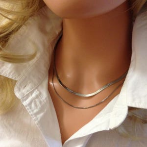May include: Two silver necklaces on a mannequin's neck. The top necklace is a flat, herringbone chain, and the bottom is a delicate box chain. The mannequin is wearing a white collared shirt.