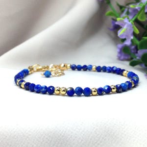 Peut inclure: Un bracelet délicat composé de petites perles bleues profondes et facettées, entrecoupées de perles dorées. Le bracelet est doté d'un fermoir doré et d'un petit charm. Le bijou est présenté sur une surface blanche avec des fleurs violettes en arrière-plan.