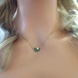 May include: A gold-coloured necklace with a delicate design. The pendant features a cluster of green and white gemstones arranged in a leaf-like pattern. The necklace is displayed on a person's neck.