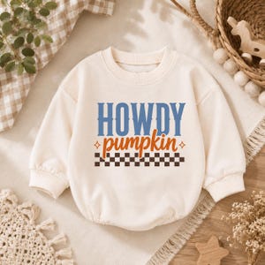 May include: A cream-colored long-sleeve sweatshirt romper featuring the text HOWDY in blue, pumpkin in orange, and a brown and white checkered pattern at the bottom. The garment has a crew neckline, ribbed cuffs, and a rounded bottom hem.