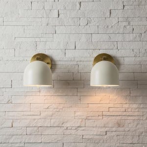 May include: Two off-white, dome-shaped wall sconces with brass-colored circular backplates are mounted on a white, textured brick wall. The sconces are illuminated, casting a warm glow downward. The design is modern and minimalist.