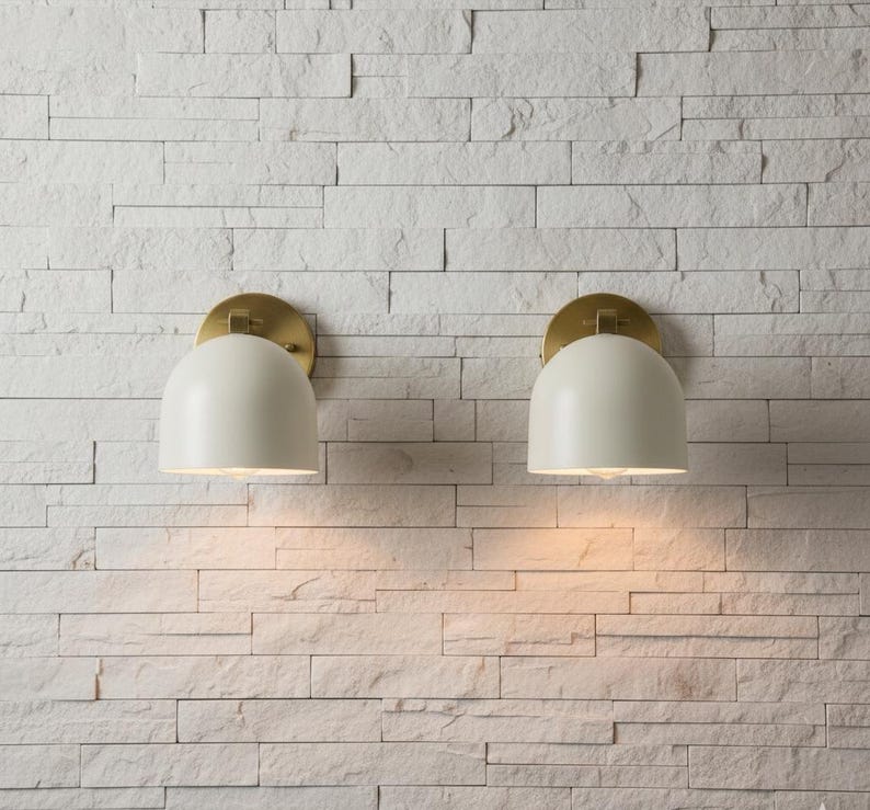 May include: Two off-white, dome-shaped wall sconces with brass-colored circular backplates are mounted on a white, textured brick wall. The sconces are illuminated, casting a warm glow downward. The design is modern and minimalist.